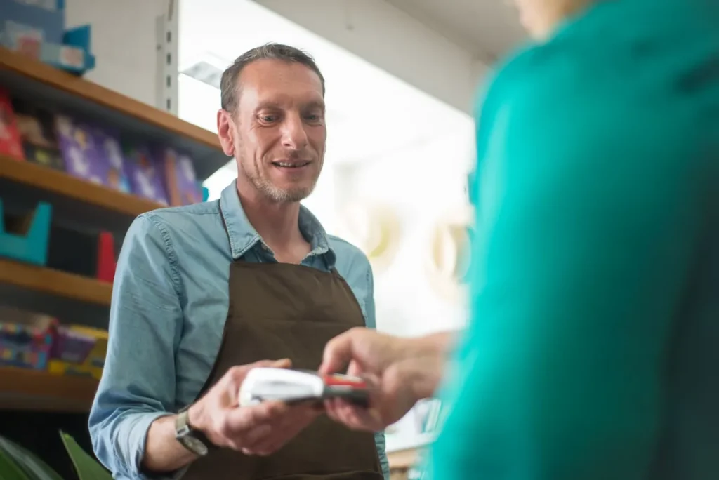 Un épicier en tablier tend un terminal de paiement à une cliente au comptoir