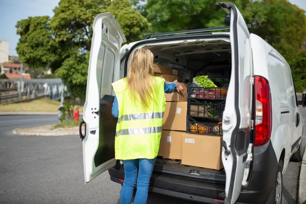 Une livreuse en gilet jaune charge son utilitaire de cartons et de caisses de légumes